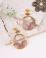 Pair of gold and purple floral earrings on a textured surface with greenery.
