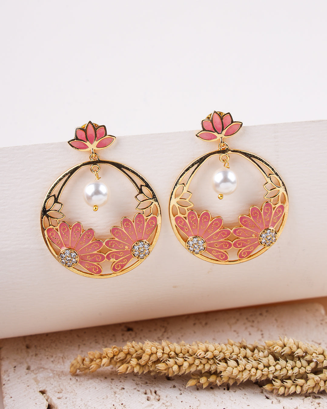 Ornate earrings with pink floral designs and pearls on a textured surface.