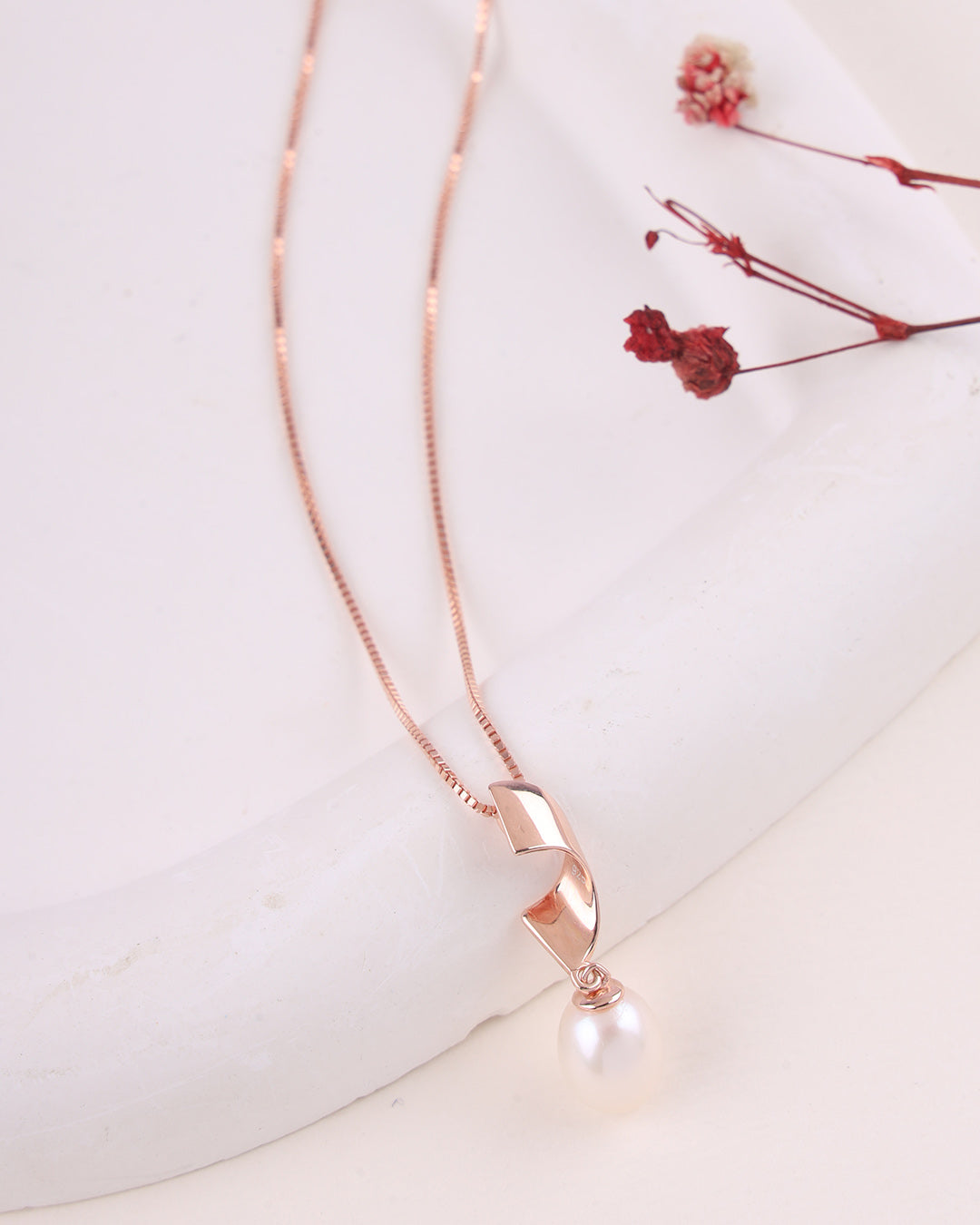 Rose gold necklace with a pearl pendant on a white surface with dried flowers.