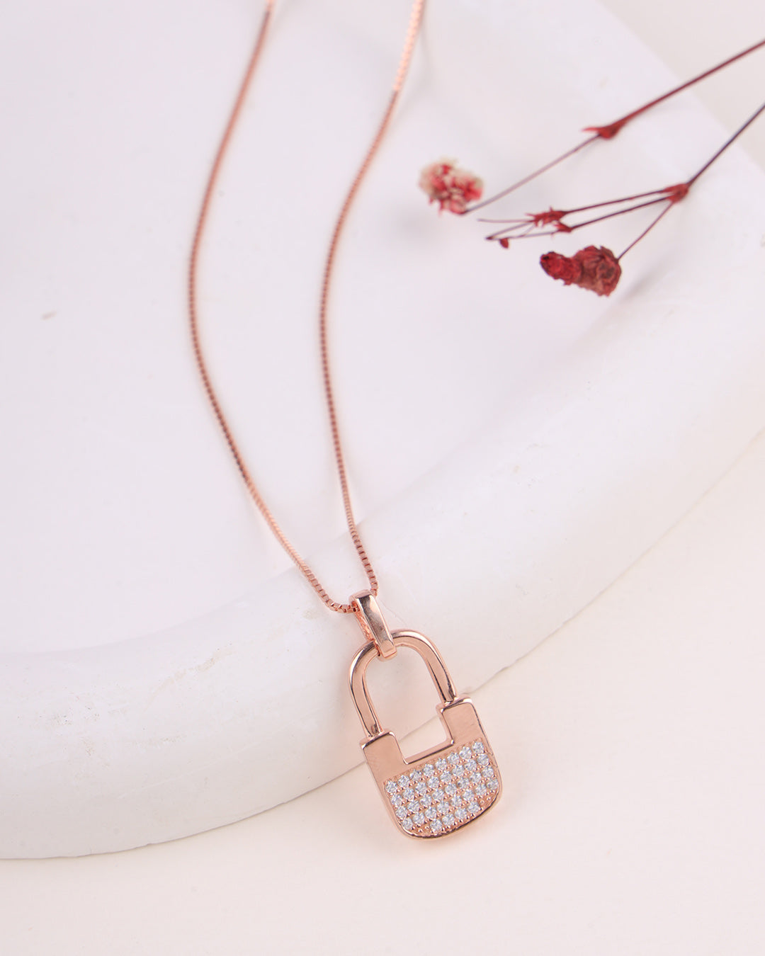 Rose gold padlock-shaped pendant with clear stones on a white surface with dried flowers.