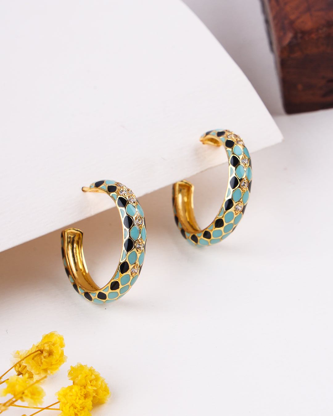 Gold hoop earrings with blue and black accents on a white surface with yellow flowers.