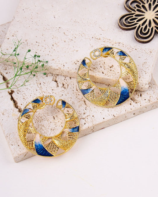 Gold and blue earrings on a textured stone surface with a small plant.