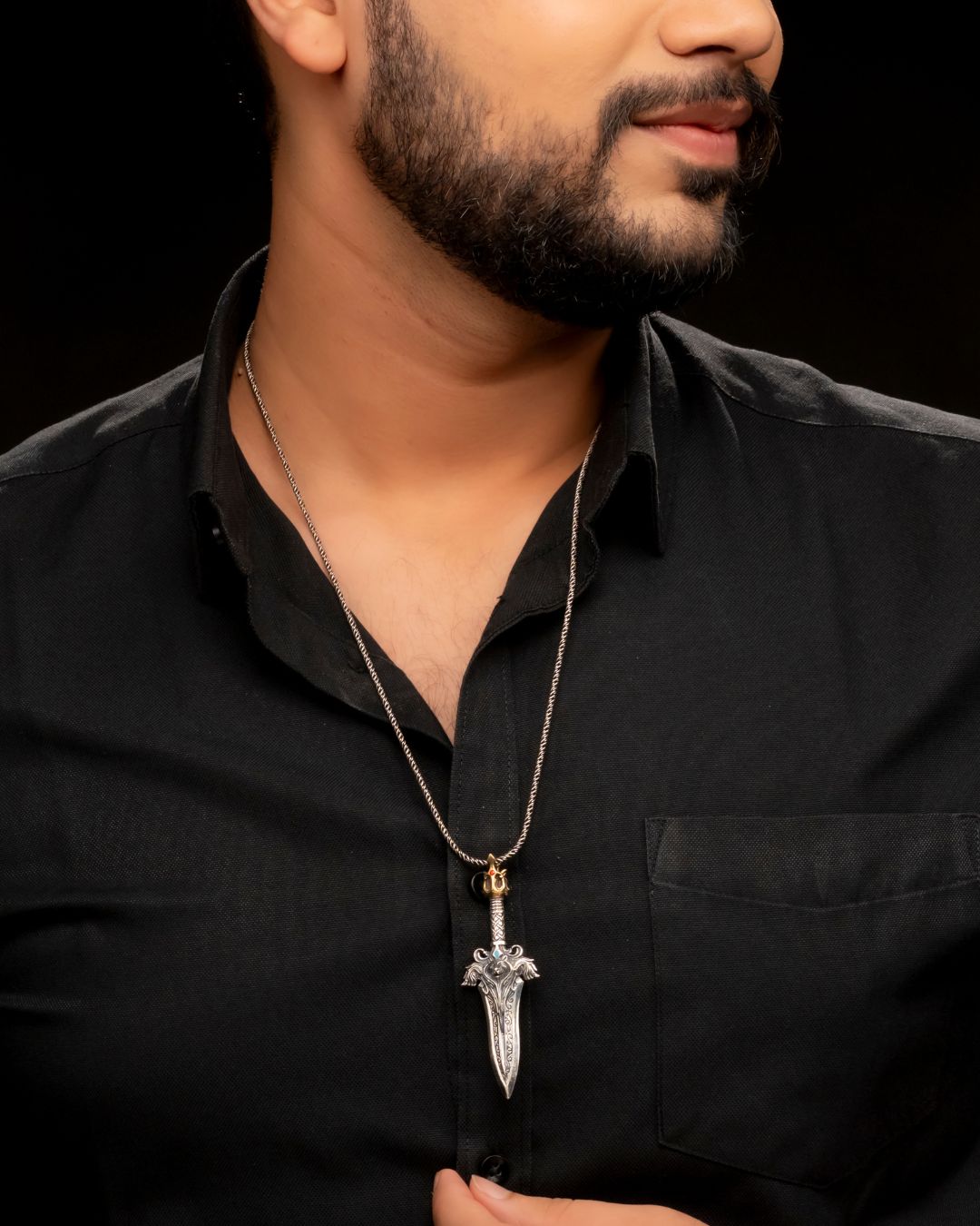 Man wearing a black shirt with a necklace featuring a dagger pendant on a dark background