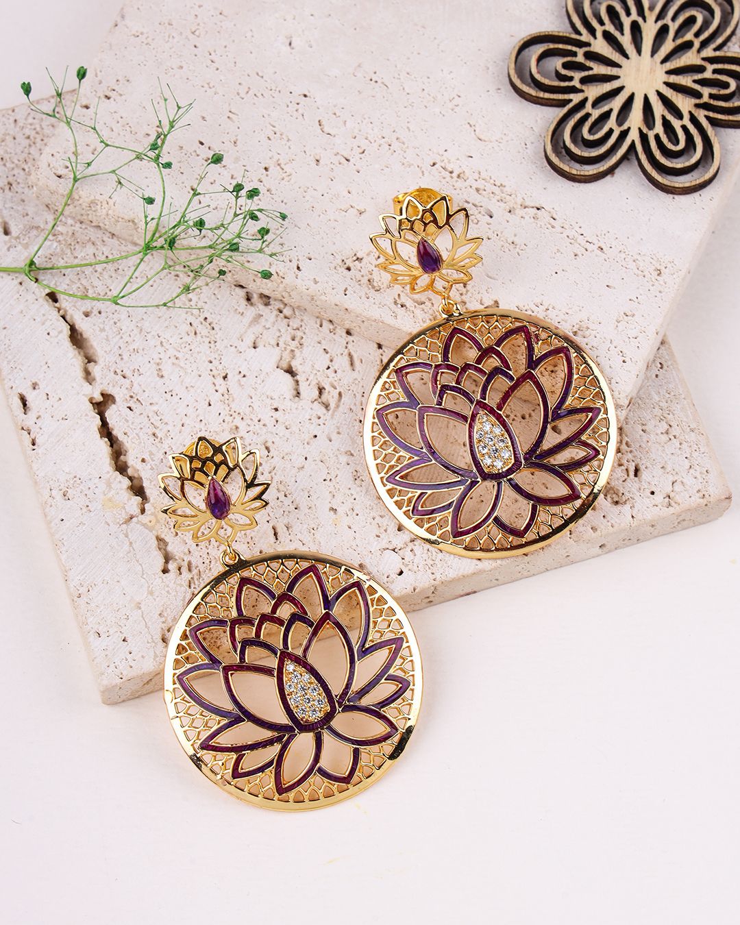 Gold and purple lotus flower earrings on a stone surface with a small plant.
