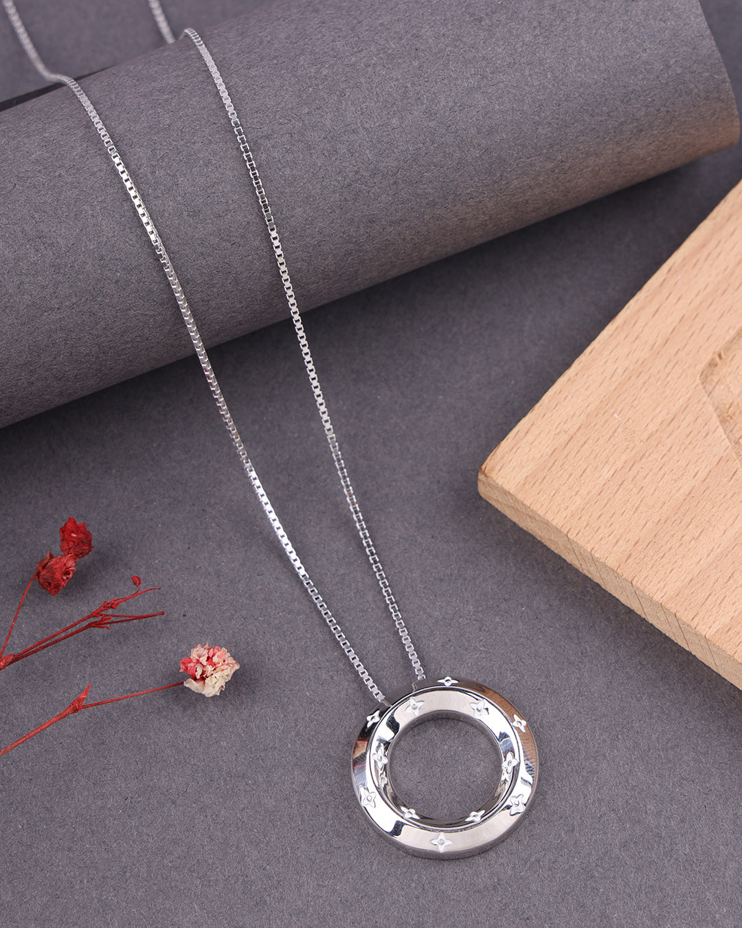 Silver necklace with a circular pendant on a gray surface with dried flowers and a wooden block.