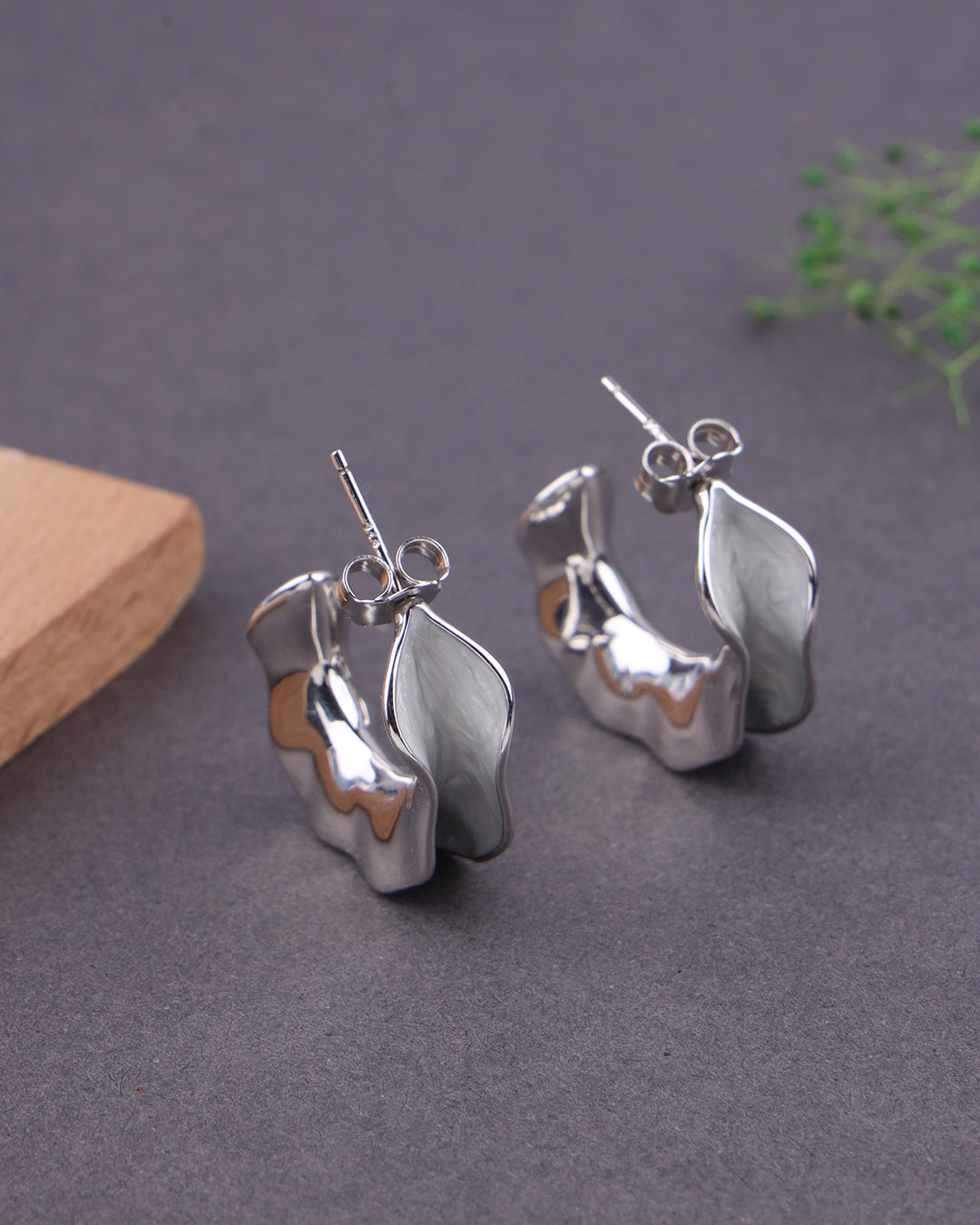 Silver leaf-shaped earrings on a gray surface with a wooden block and green leaves in the background.