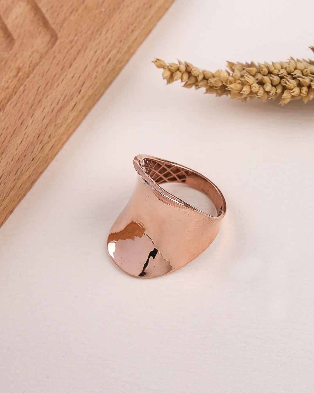 Rose gold ring on a light surface with a wooden board and dried plants in the background