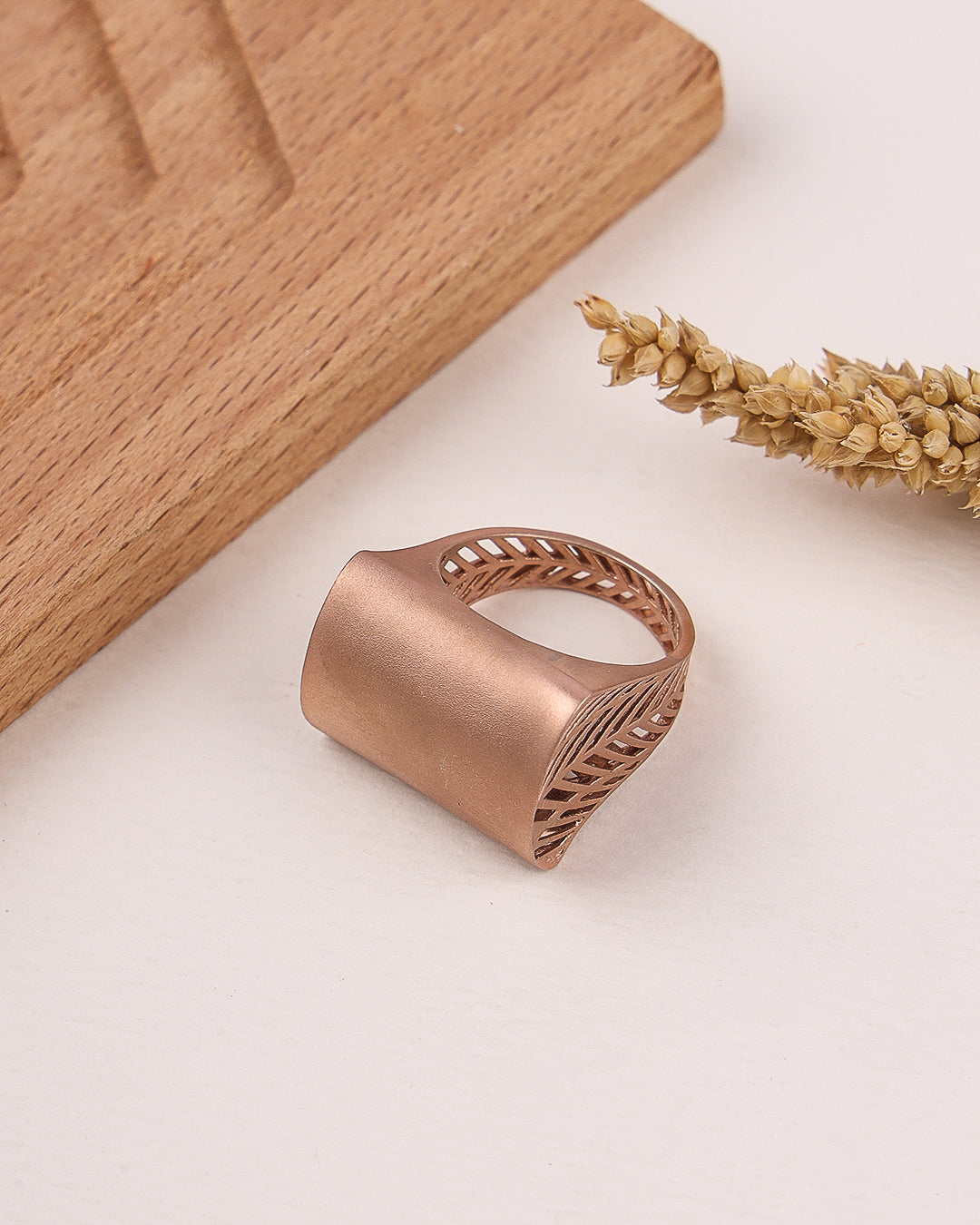 Rose gold ring with intricate design on a light surface with wooden block and dried plant.