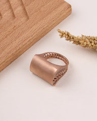 Rose gold ring with intricate design on a light surface with wooden block and dried plant.