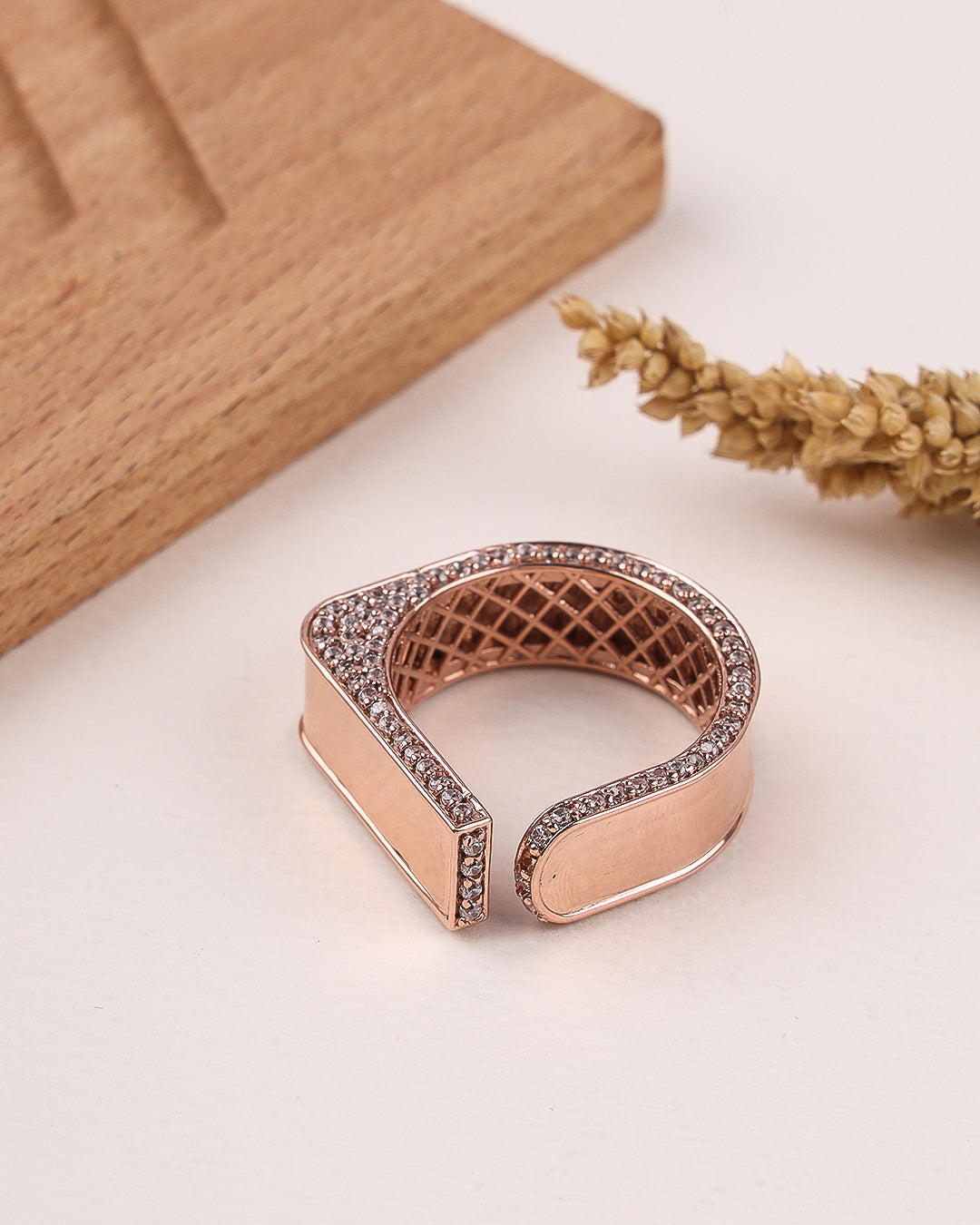 Rose gold ring with diamond accents on a light surface with a wooden block and dried flowers.