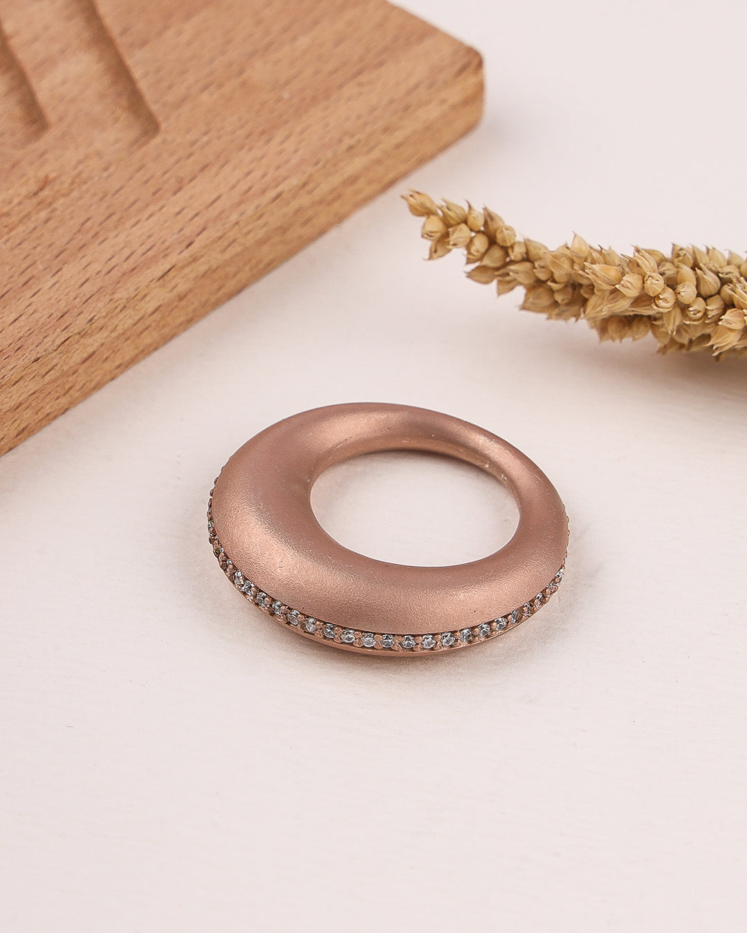 Rose gold ring with embedded stones on a light surface with wooden block and dried plant.