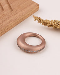 Rose gold ring with embedded stones on a light surface with wooden block and dried plant.
