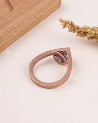 Rose gold ring with a purple gemstone on a light surface with wooden block and dried flowers.