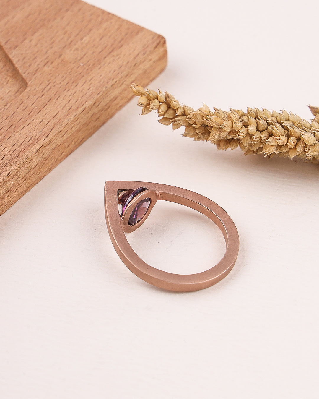 Rose gold ring with a purple gemstone on a light surface with wooden block and dried plant.