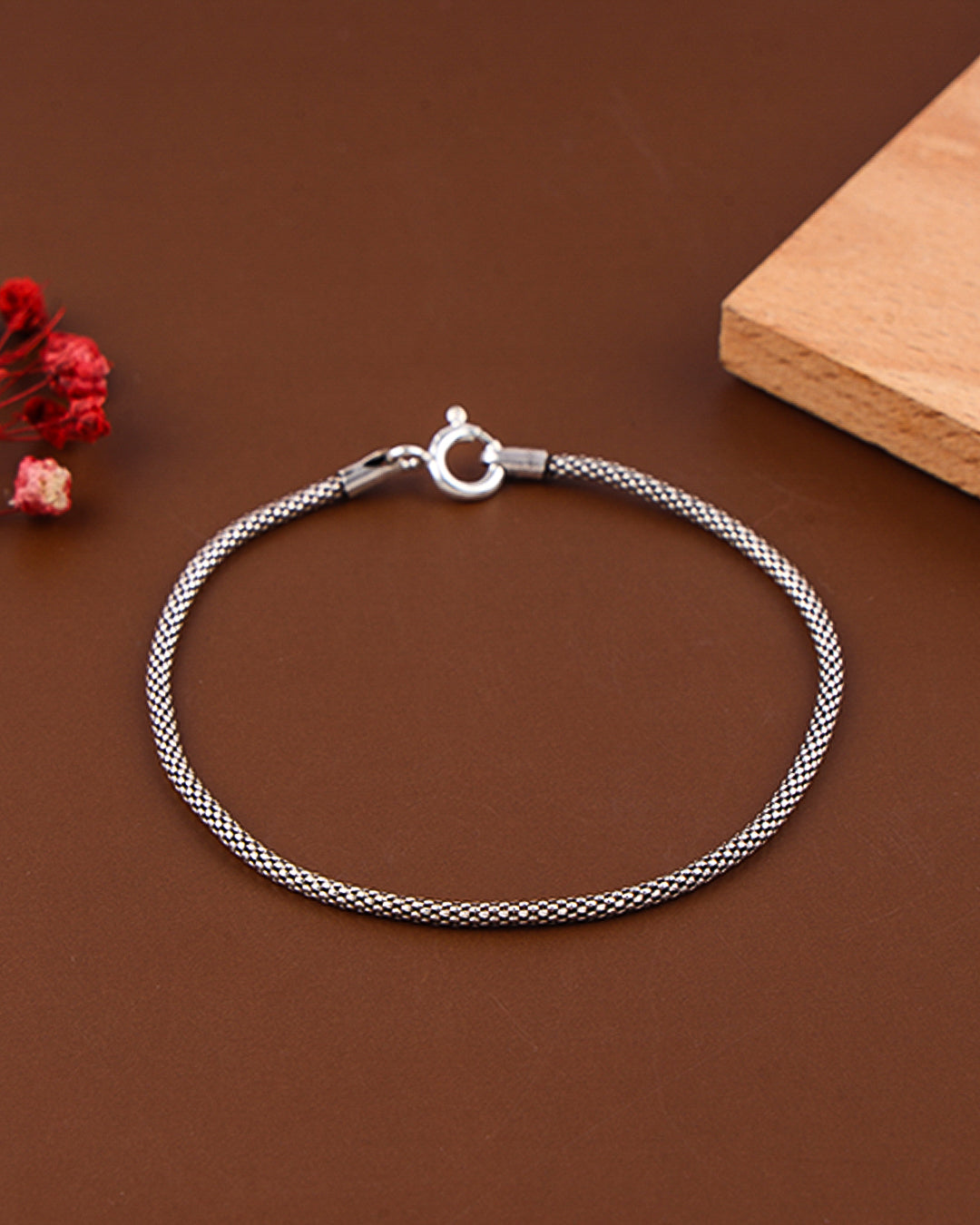 Silver braided bracelet on a brown surface with a wooden block and red flowers.