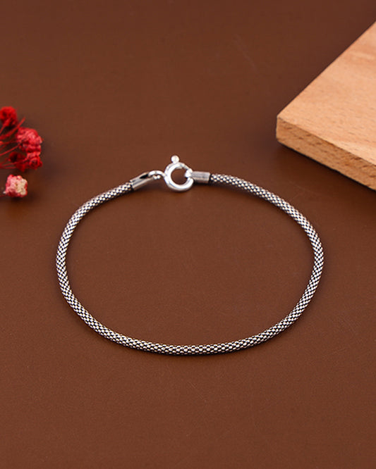 Silver braided bracelet on a brown surface with a wooden block and red flowers.