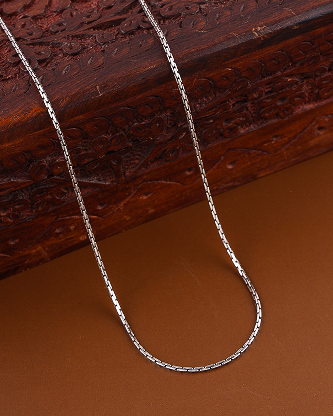 Silver chain necklace on a brown surface with a wooden box in the background