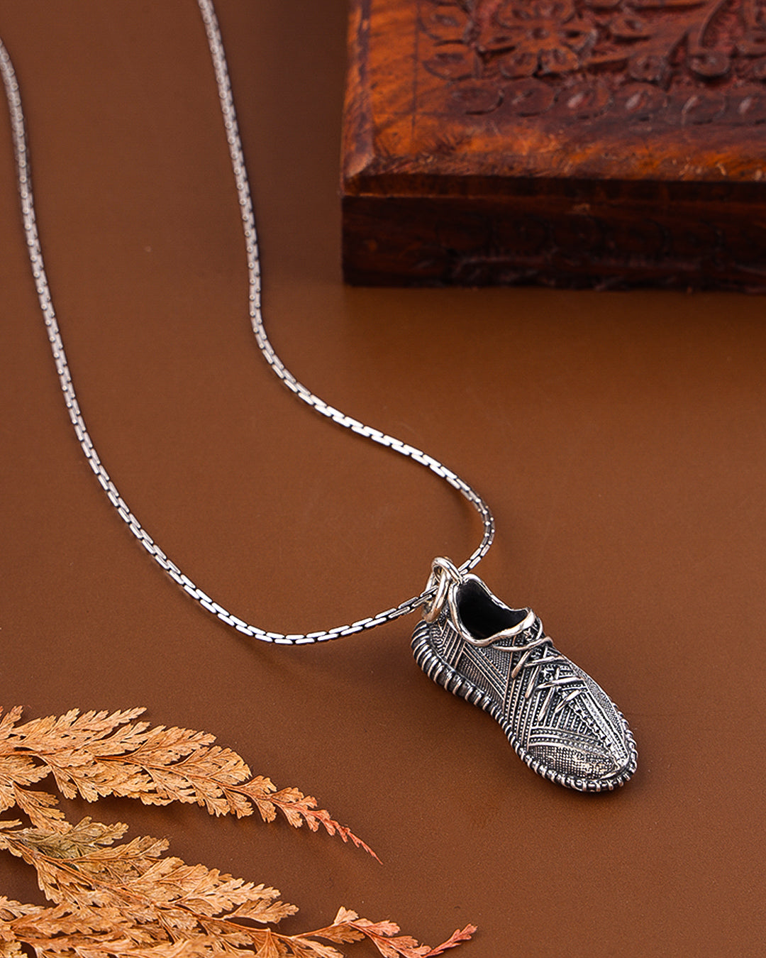 Silver shoe-shaped pendant on a chain against a brown background with a wooden block and dried leaves.
