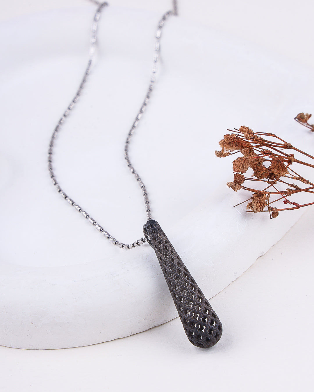 Necklace with a textured pendant on a white surface with dried flowers.