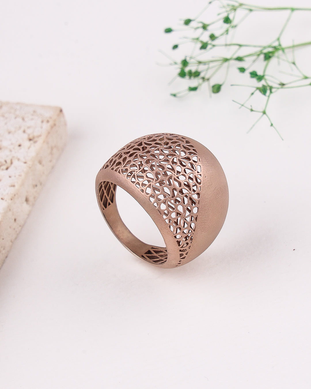 Rose gold ring with intricate design on a white surface with a natural stone and greenery.