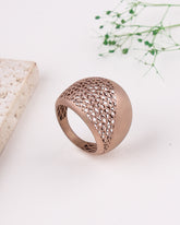 Rose gold ring with intricate design on a white surface with a natural stone and greenery.