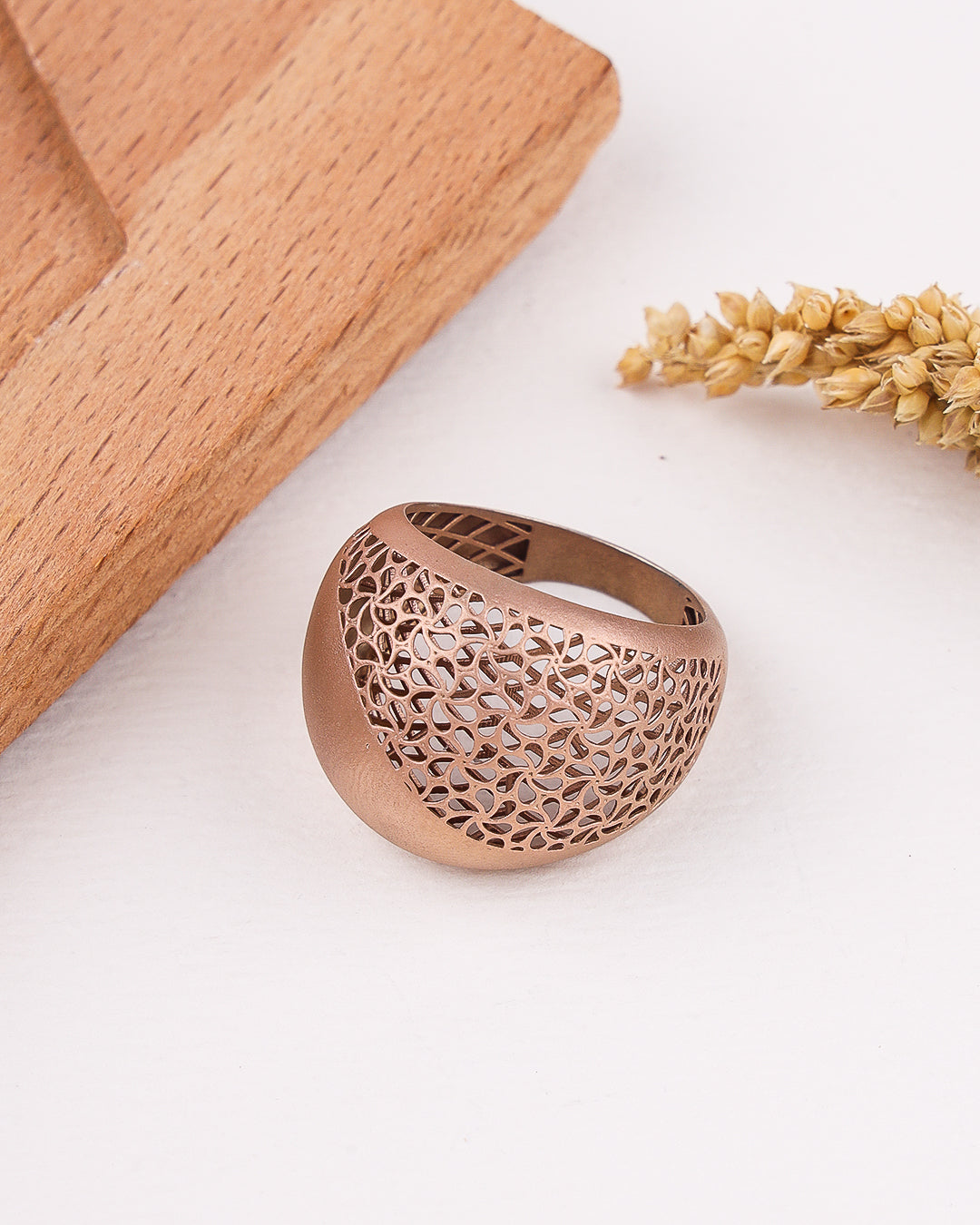 matte rose gold ring with intricate design on a light surface with wooden block and dried plants.