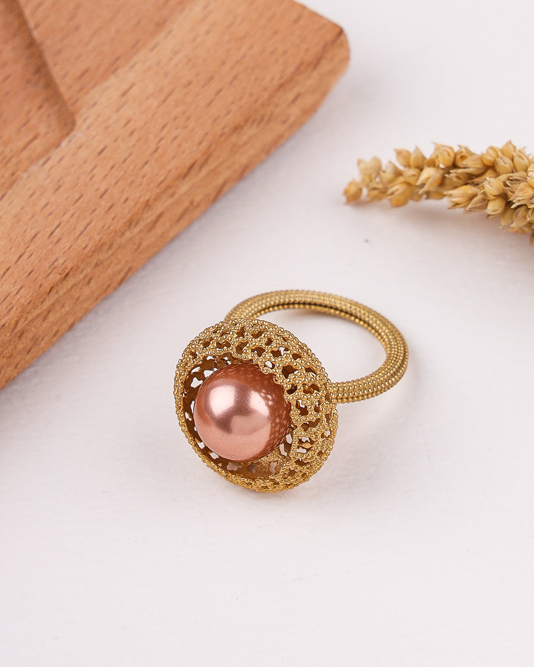 Gold ring tie with a pink pearl on a white surface with wooden block and dried plant