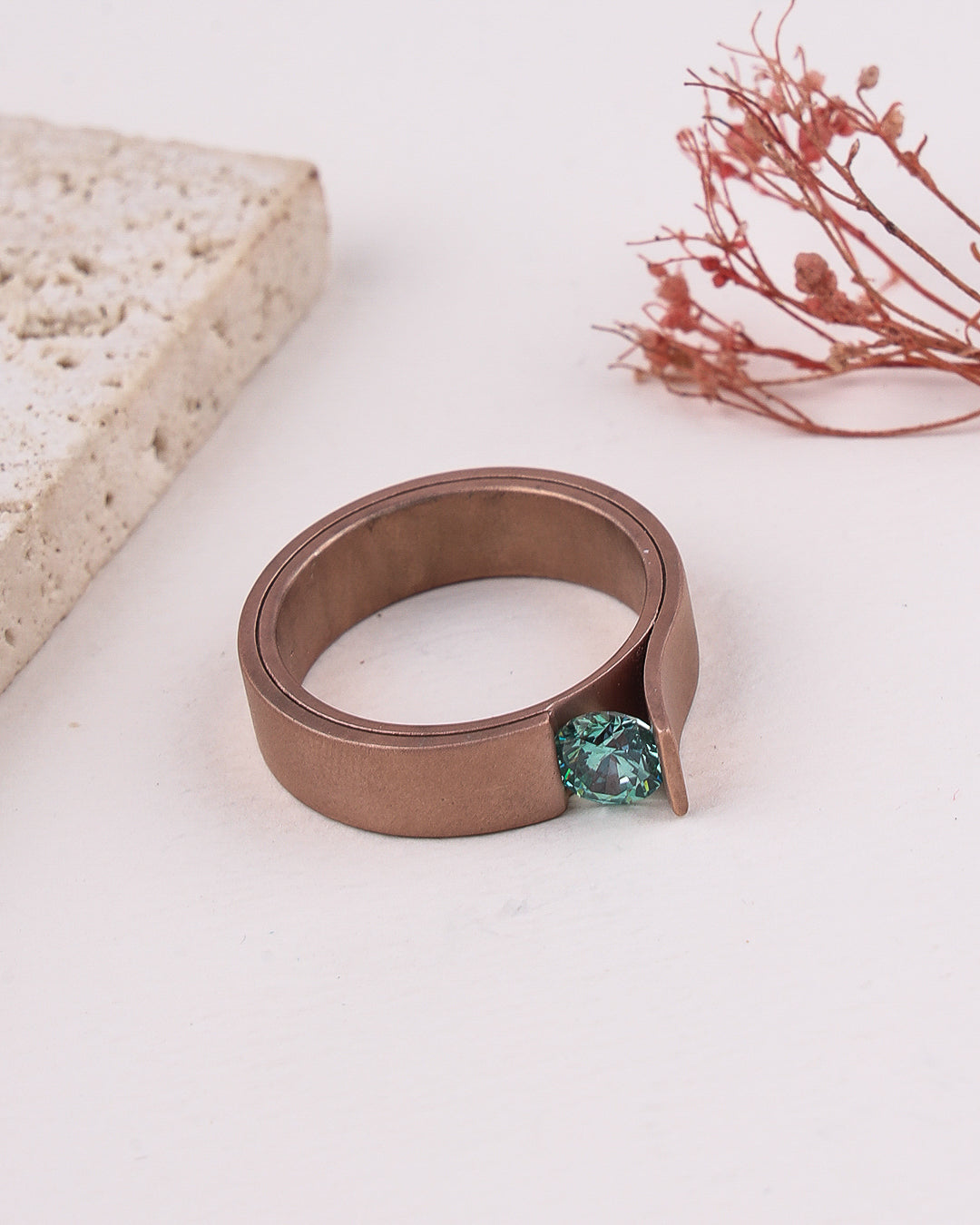matte rose gold ring with a green gemstone on a white surface with dried flowers and stone in the background
