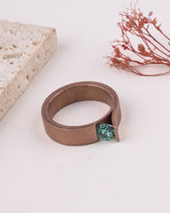 matte rose gold ring with a green gemstone on a white surface with dried flowers and stone in the background