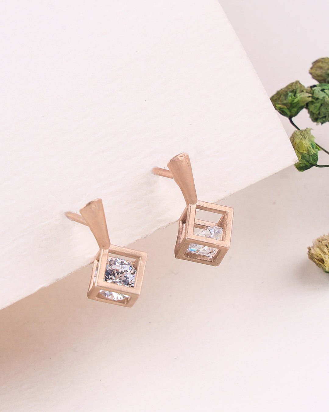Rose gold earrings with clear gemstones on a light surface with greenery.