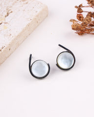 Black rhodium earrings on a white surface with a textured beige object and dried flowers in the background.