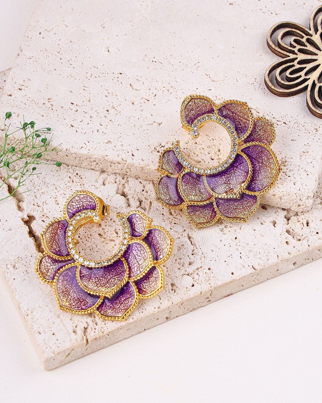 Purple and gold earrings on a textured stone surface with a plant and wooden piece.