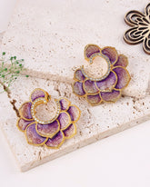 Purple and gold earrings on a textured stone surface with a plant and wooden piece.