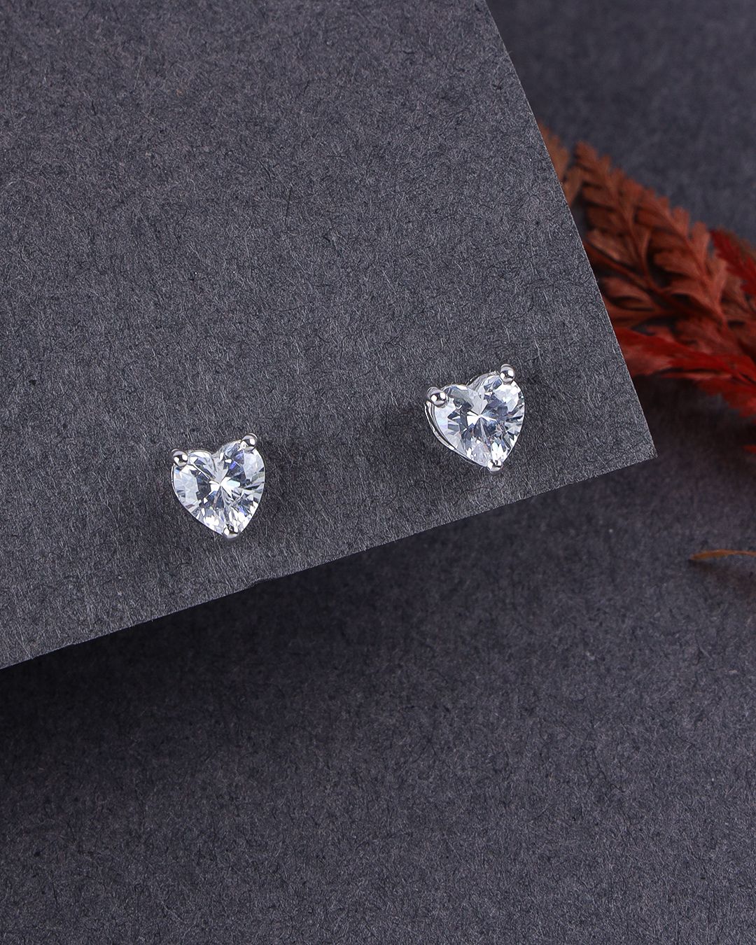 Heart-shaped stud earrings on a dark gray surface with a blurred red flower in the background.