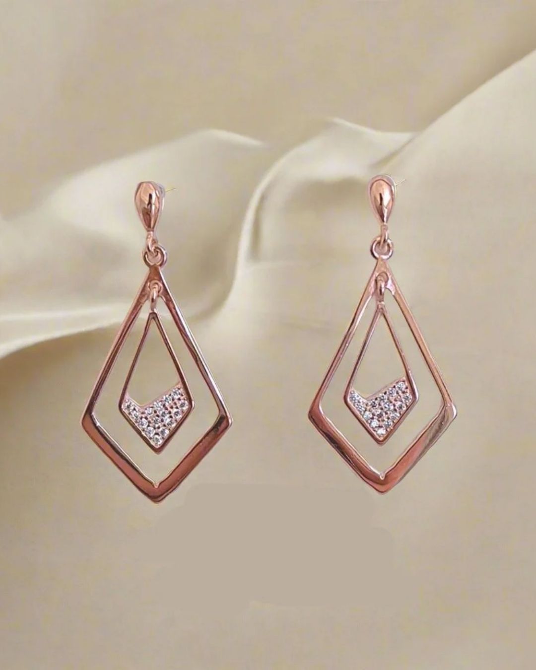 Rose gold earrings with diamond-like stones on a beige fabric background