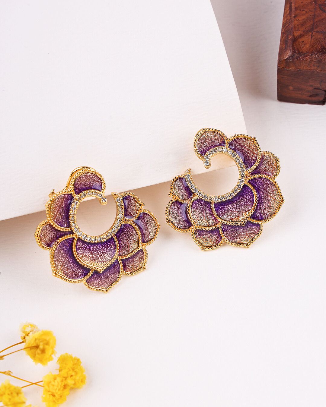 Purple floral earrings with gold accents on a white surface with yellow flowers and a wooden box.