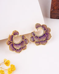 Purple floral earrings with gold accents on a white surface with yellow flowers and a wooden box.
