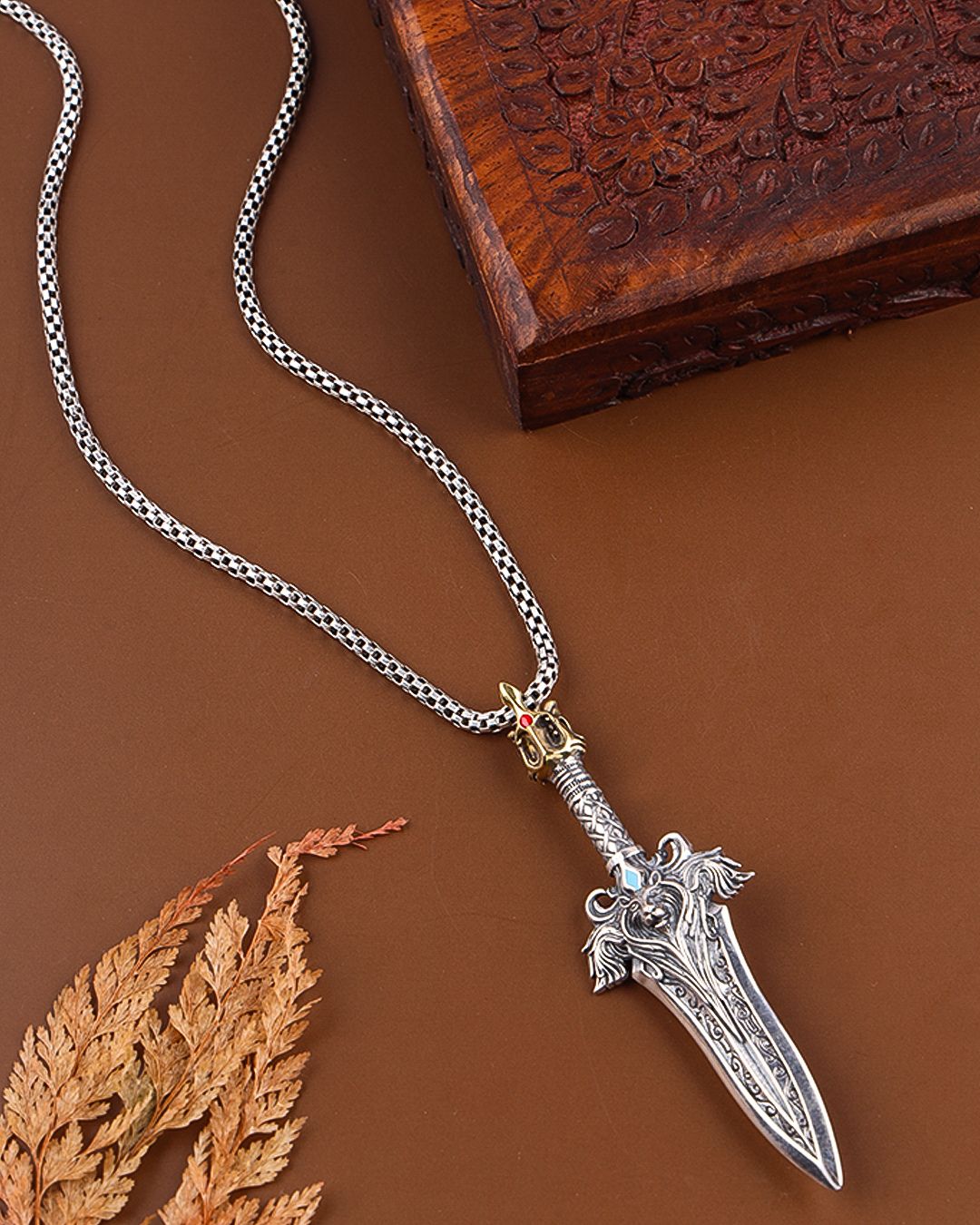 Silver dagger-shaped pendant on a chain with a wooden box and leaf in the background