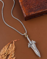 Silver dagger-shaped pendant on a chain with a wooden box and leaf in the background