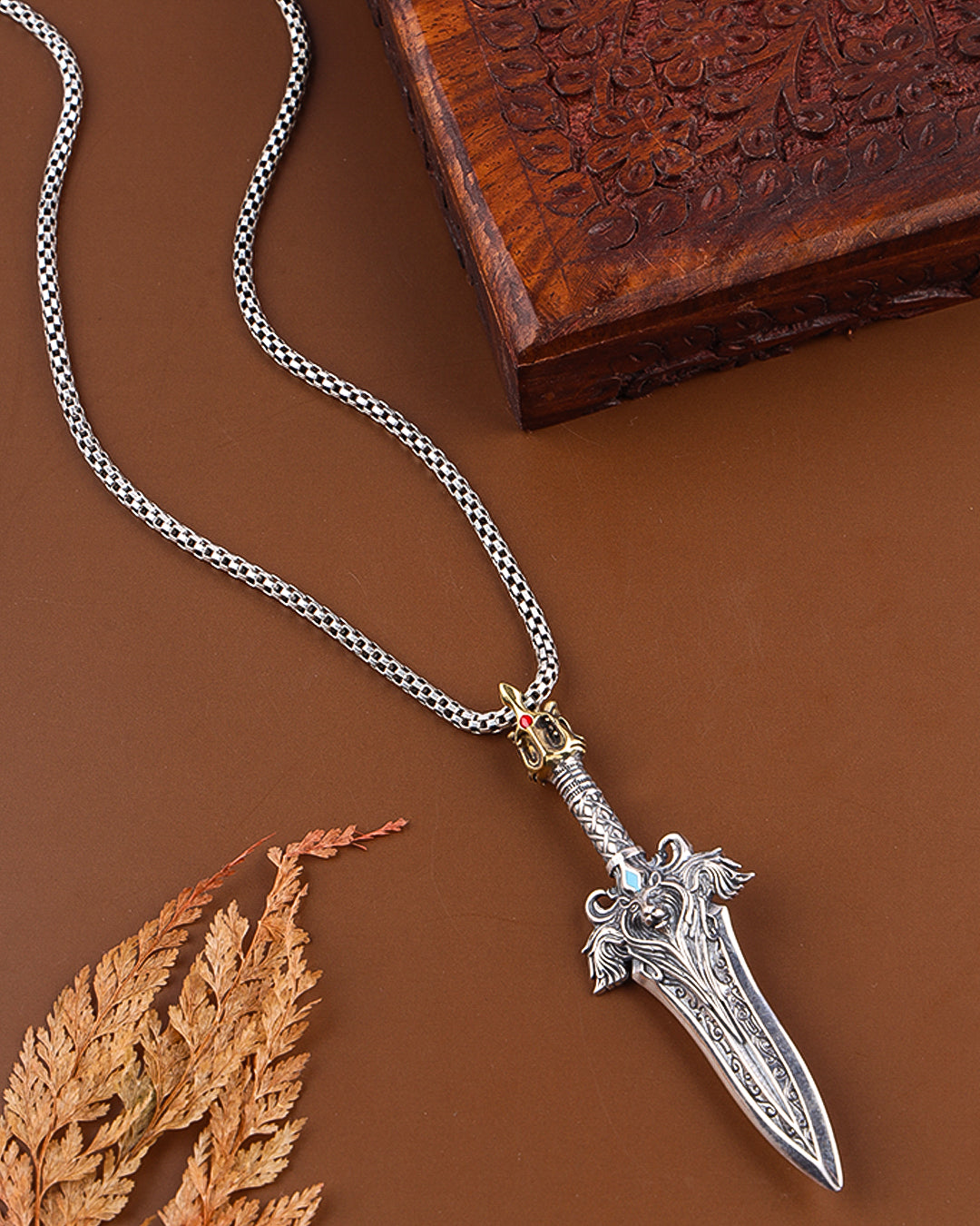 Silver dagger pendant necklace on a brown background with a wooden box and leaf.