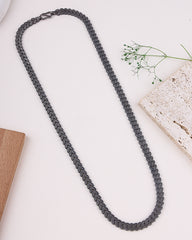 Black rhodium necklace on a white surface with a wooden block and plant in the background