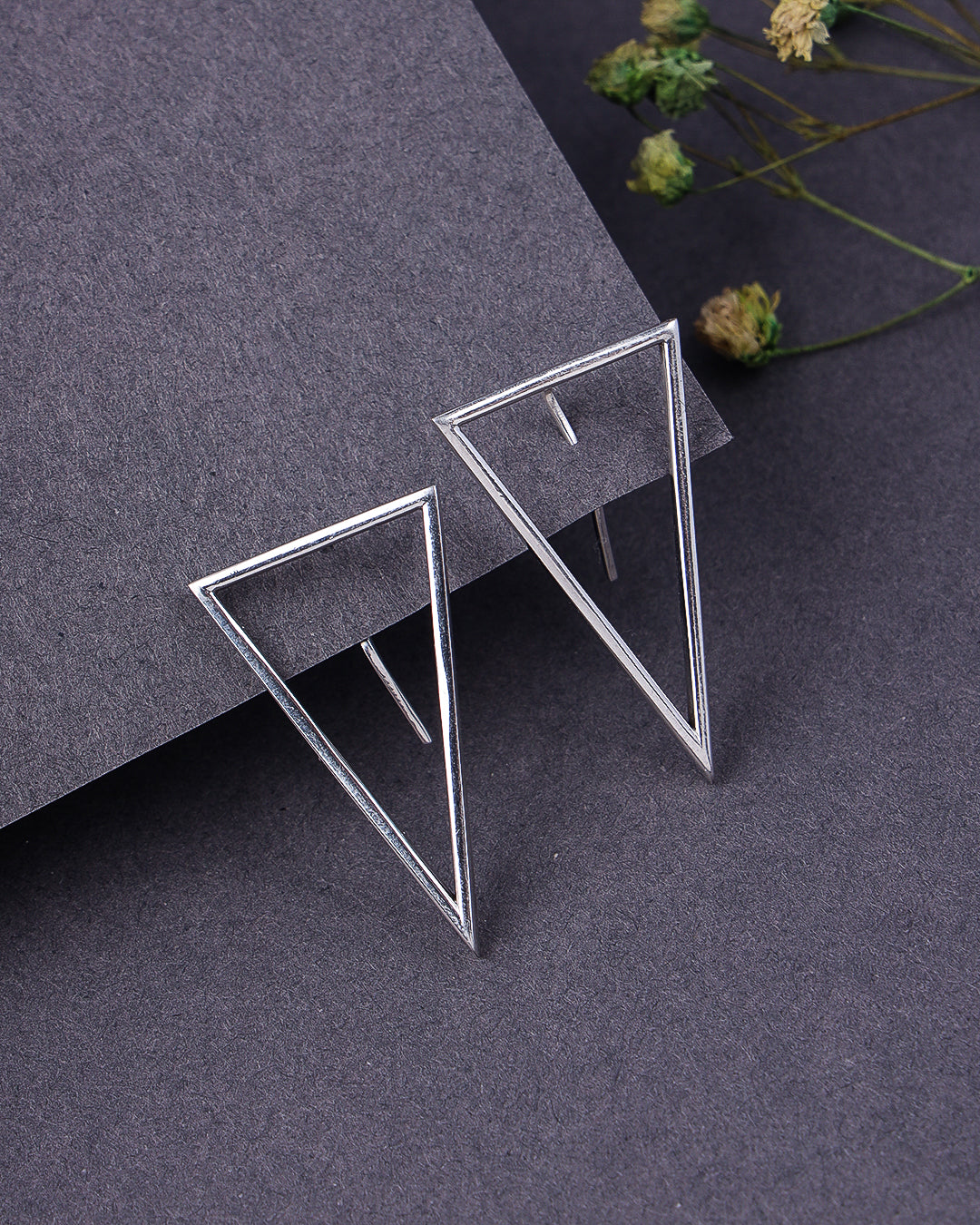 Silver triangle earrings on a dark gray surface with dried flowers.