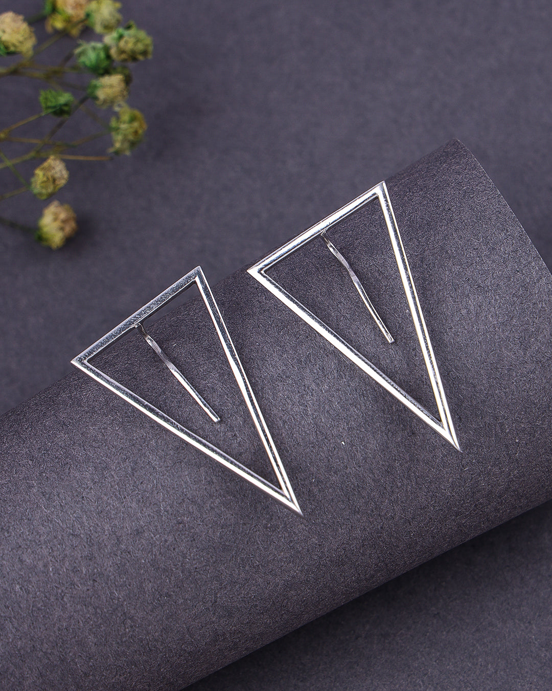 Silver geometric earrings on a dark textured surface with dried flowers.