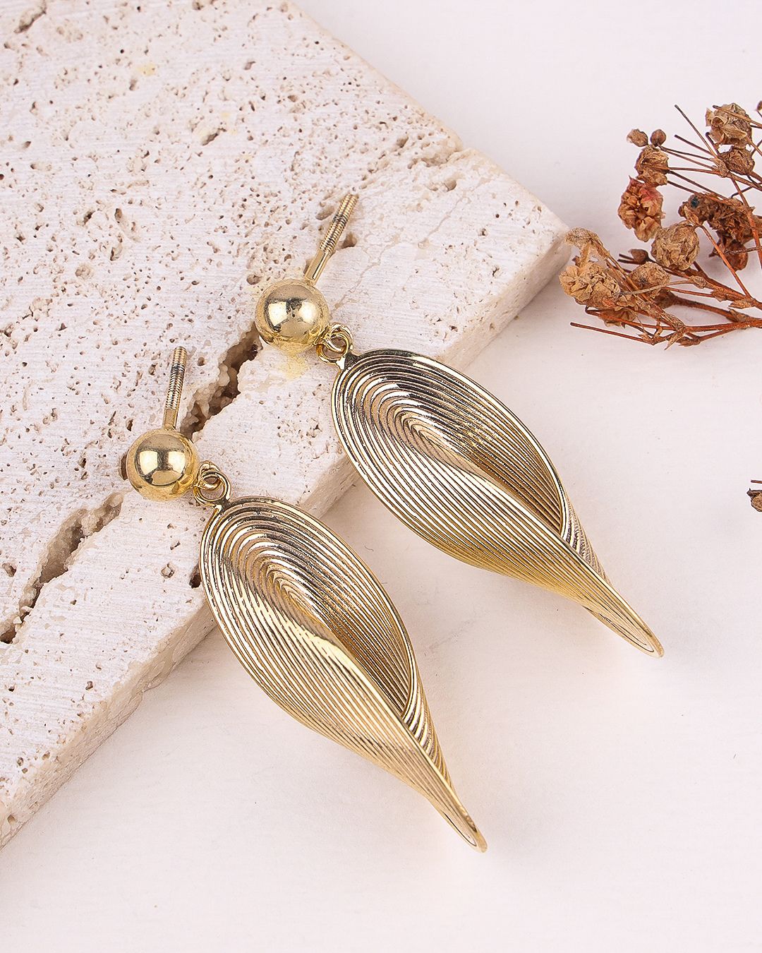 Gold leaf-shaped earrings on a textured surface with dried flowers.
