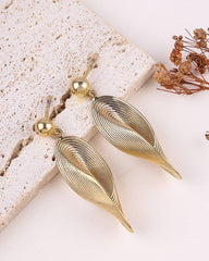Gold leaf-shaped earrings on a textured surface with dried flowers.