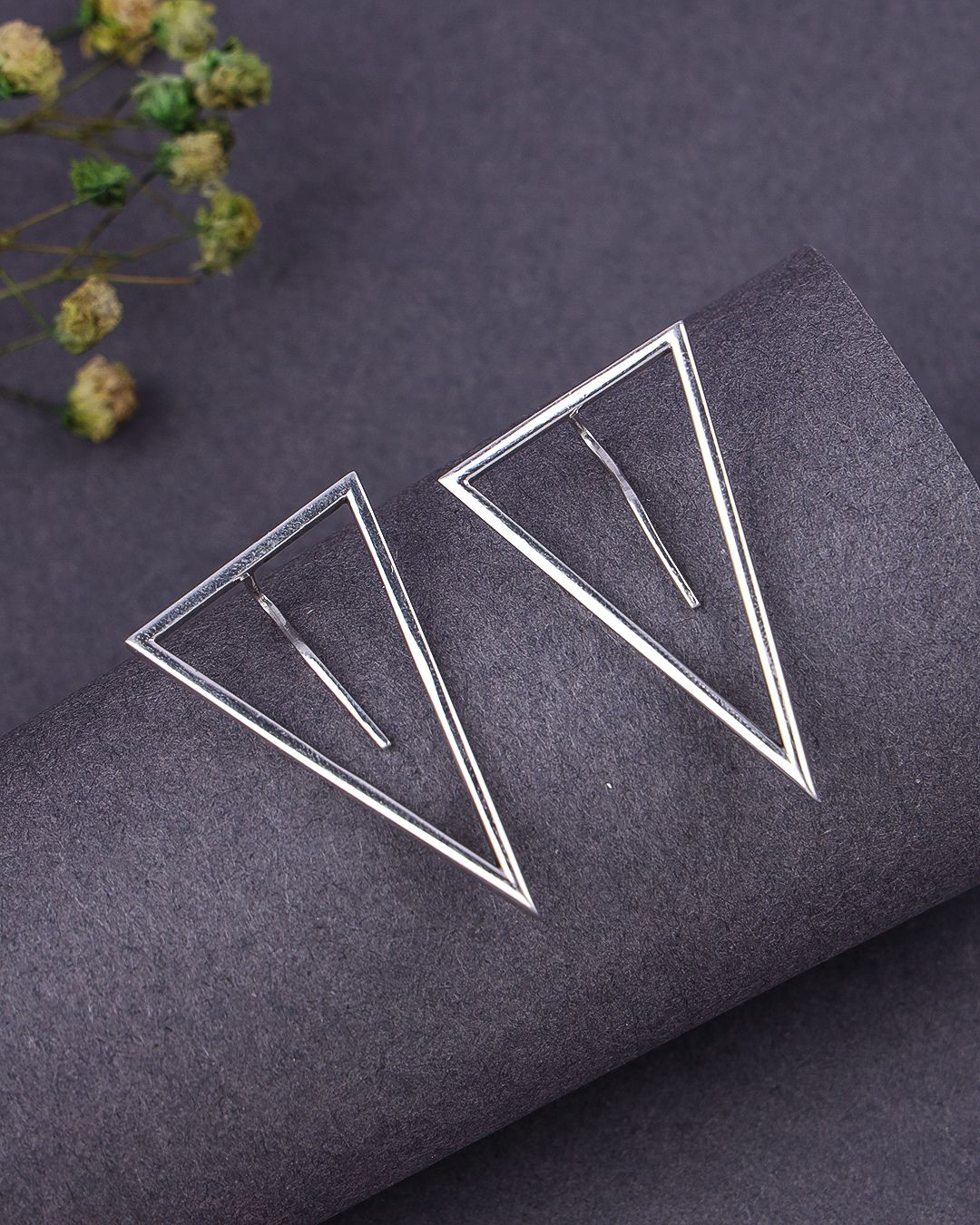 Pair of geometric silver earrings on a dark textured surface with dried flowers.
