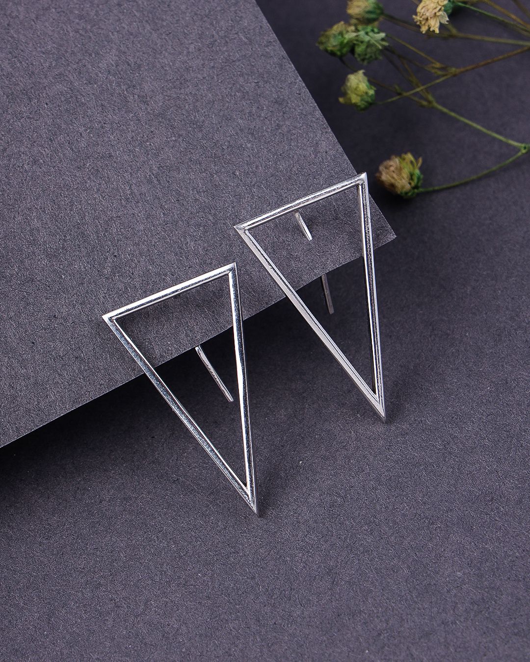 Pair of geometric silver earrings on a dark gray surface with dried flowers.
