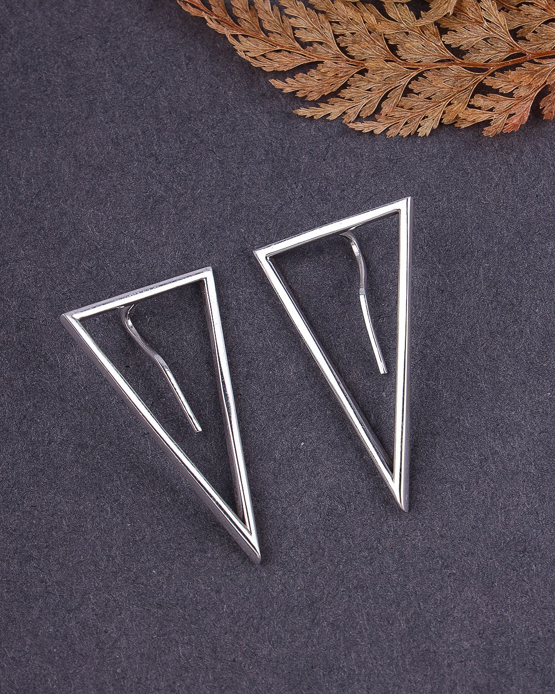 Pair of silver triangle earrings on a dark surface with a leaf in the background