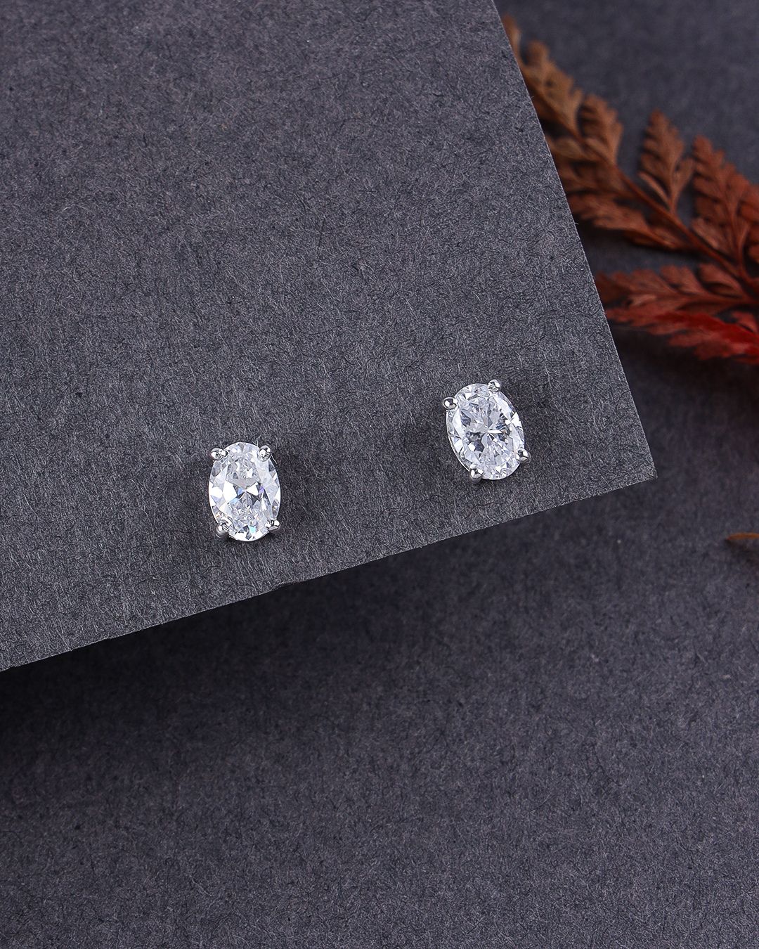 Pair of silver oval stud earrings on a dark gray surface with dried leaves.