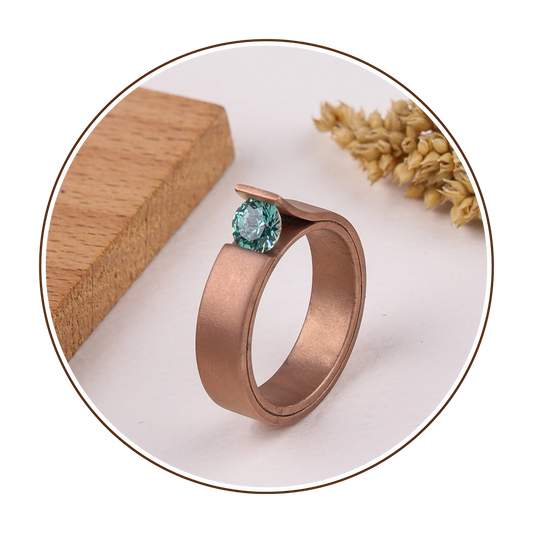 Rose gold ring with a green gemstone on a white surface with wooden and dried plant elements.